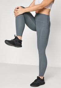 Leggings gris ajustés avec des accents de couture et des poches latérales, associés à des chaussures de sport noires, présentés en position debout.