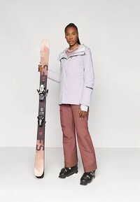 Γυναίκα με μωβ μπουφάν και μωβ παντελόνι κρατάει μια σανίδα του σκι όρθια, φορώντας μαύρες μπότες του σκι, στέκεται σε λευκό φόντο.