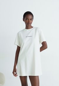 Balta didelė t-shirt suknelė, pagaminta iš minkštos medvilnės, su trumpomis rankovėmis ir apvalia iškirpte; priekyje turi juodą užrašą.