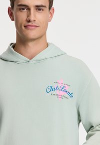 Mintovo zelená mikina s kapucňou s jemnou textúrou, s viacfarebnou potlačou loga: "Club Locale" s ružovými a modrými akcentmi.