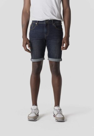 Hombre con pantalones cortos de mezclilla oscuros enrollados, zapatillas blancas y camiseta blanca, de pie contra un fondo claro y liso.