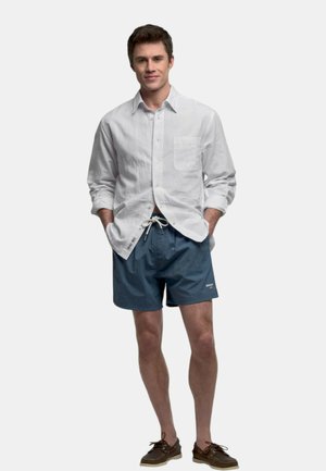 Uomo in piedi con le mani nelle tasche che indossa una camicia bianca a maniche lunghe, pantaloncini blu con coulisse e mocassini marroni su sfondo chiaro.