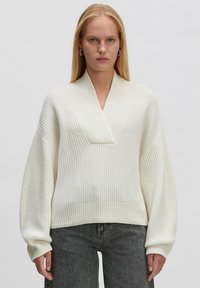 Helles Creme gestricktes Sweatshirt mit V-Ausschnitt, übergroßen Ärmeln, geripptem Saum und strukturiertem Muster; kombiniert mit dunkelgrauen Jeans.