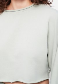 Gros plan sur le torse portant un haut à manches longues raccourci gris clair avec un ourlet à bord brut et des coutures visibles au col.