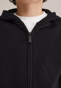 Melnā kapucēm pieguļoša svetkrekls, izgatavots no kokvilnas un sintētisko šķiedru maisījuma, ar paaugstinātu apkakli, priekšējo rāvējslēdzēju un kapuci, ar minimāliem šuvju detaļām.