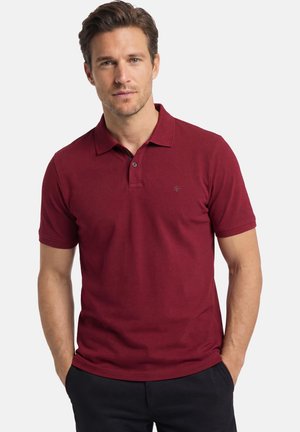 Mann mit hellem Teint und braunen Haaren, der ein dunkelrotes Poloshirt mit kurzen Ärmeln trägt und mit den Händen in den Taschen vor weißem Hintergrund steht.