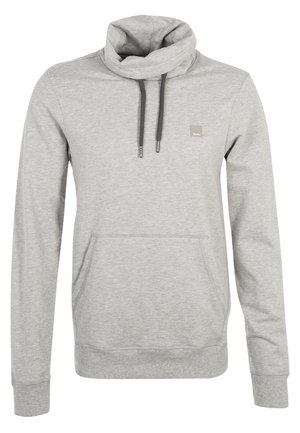 Homme portant un sweat à capuche gris, un pantalon noir et des baskets bordeaux, portant une veste grise sur l'épaule en marchant vers l'avant.