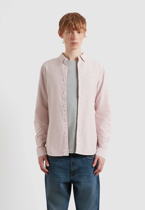 Giovane con capelli rossi corti che indossa una camicia a righe rosa e bianca sopra una t-shirt grigia e jeans blu, in posizione neutra.