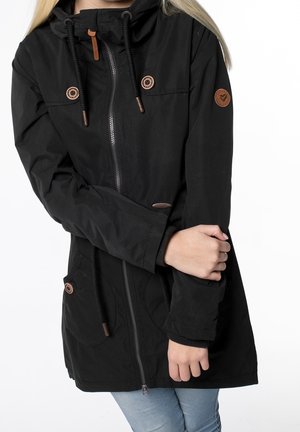 Mujer con chaqueta negra con capucha, cremallera frontal, botones decorativos y un emblema de corazón en la manga izquierda, combinada con jeans azul claro.