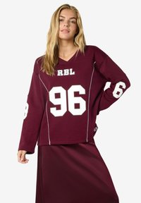Bordo oversized sportski dres s bijelim detaljima i brojem "96" otisnutim na prednjem dijelu i rukavima; mekani materijal s V-izrezom.