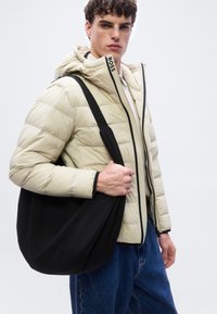 Νεαρός άνδρας που φοράει εκρού μπουφάν με κεντήματα και το λογότυπο "BOSS", μπλε τζιν, κρατώντας μεγάλη μαύρη τσάντα ώμου μπροστά σε απλό φόντο.