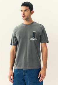 Graues kurzärmeliges T-Shirt mit einem Guinness-Logo-Grafik auf der linken Brust und einem lockeren Schnitt, kombiniert mit blauen Jeans.