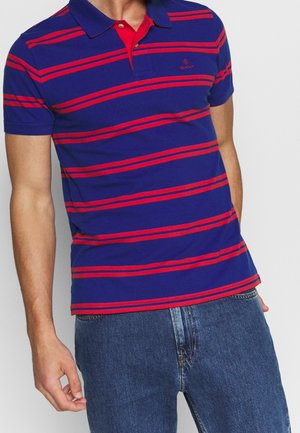 Marineblaue Poloshirt mit roten horizontalen Streifen, ausgestattet mit einem roten Kragen, dezentem Logo-Stick und kurzen Ärmeln. Kombiniert mit Jeans.