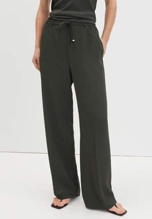 Woman wearing loose dark green wide-leg pants with drawstring waist, paired with black sleeveless top and black open-toe sandals.