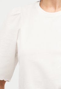 Λευκό τοπ με φουσκωτά μανίκια με κυματιστά άκρα στα μανίκια, στρογγυλή λαιμόκοψη και υφασμάτινη υφή στο πάνω μέρος του σώματος ενός ατόμου.