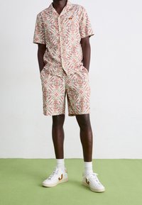 Ensemble chemise à manches courtes et short à motif floral, aux couleurs douces de pêche, crème et vert. Baskets blanches avec des accents marron.