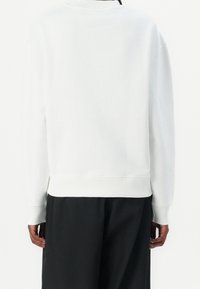 Personne portant une chemise blanche unie à manches longues et un pantalon noir, vue de dos sur fond blanc.