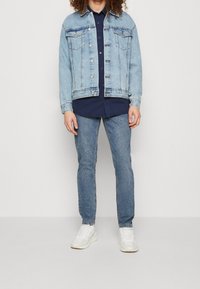 Chaqueta de denim azul claro con dos bolsillos en el pecho, cierre de botones y yugo trasero. Lleva puesta sobre una camisa azul oscura, combinada con jeans azules y zapatillas blancas.