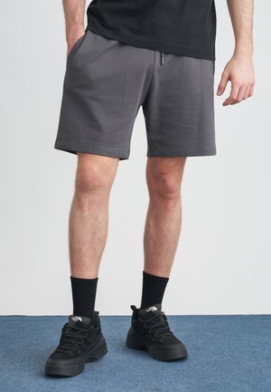 Γκρι αθλητικό σορτς με ελαστική μέση, πλαϊνές τσέπες και λεία υφή. Συνδυασμένο με μαύρα αθλητικά παπούτσια και κάλτσες μήκους μέχρι τον αστράγαλο.