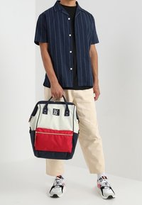 Plecak z płótna w kolorach granatowym, kremowym i czerwonym. Posiada podwójne uchwyty, zewnętrzną kieszeń na zamek i naszywkę z logo. Strukturalny design o kątnym kształcie.