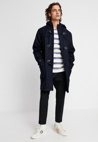 Cappotto duffle in lana blu navy con chiusura a toggles, grandi tasche e cappuccio, indossato sopra una t-shirt a righe con scollo a barca e pantaloni scuri.