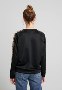 Svart sweatshirt med en slät textur, försedda med gulddetaljer längs axlarna och raglanärmarna. Kombinerad med blå denimjeans.