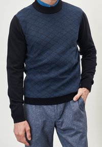 Uomo che indossa un maglione blu a fantasia texture con maniche nere e pantaloni blu a texture, con una mano in tasca.