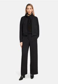 Czarna, krótka kurtka zapinana na zamek z wysokim kołnierzem, zestawiona z czarnymi, szerokimi spodniami oraz czarnymi botkami na obcasie. Tkanina wydaje się miękka i o wyraźnej fakturze.