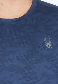 Námorně modrá košile s krátkým rukávem se jemným maskáčovým vzorem, vyrobená z texturované tkaniny, s stříbrným logem pavouka na levé straně.