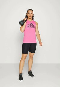Maglietta atletica rosa senza maniche con logo nero, pantaloni corti neri e scarpe sportive nere. In mano una kettlebell con impugnatura testurizzata.