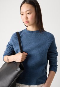 Blauwe gebreide trui met geribbelde halslijn en manchetten, gedragen met een zwarte leren tote bag. De textuur is zacht en licht wollig.