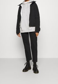 Svart jacka med huva över en ljusgrå huvtröja, tillsammans med mörka byxor och svarta snörkängor. Texturerade material och enkel design.