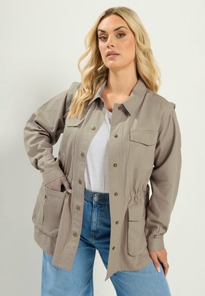 Kobieta o długich blond włosach, ubrana w beżową zapinaną na guziki kurtkę na białą koszulę i niebieskie dżinsy, z jedną ręką w kieszeni kurtki.