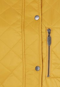 Gele gewatteerde jas met ruitpatroon, zilveren drukknoopjes en een contrasterende rits met metalen trekker. Gave textuur.