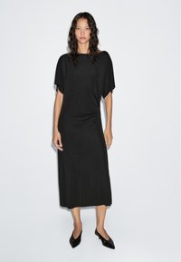 Schwarzes Midikleid mit kurzen Fledermausärmeln, aus weichem Stoff gefertigt. Verfügt über einen abgerundeten Saum und eine lässige Passform, kombiniert mit schwarzen spitzen Schuhen.