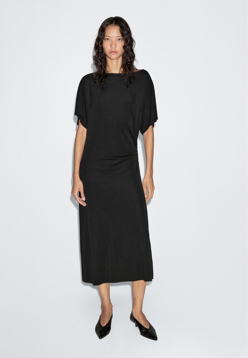 Schwarzes Midikleid mit kurzen Fledermausärmeln, aus weichem Stoff gefertigt. Verfügt über einen abgerundeten Saum und eine lässige Passform, kombiniert mit schwarzen spitzen Schuhen.