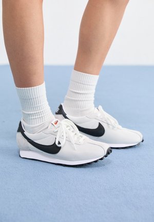 Osoba stojící na světle modrém koberci, obutá v bílých teniskách Nike s černým logem Swoosh a bílých ponožkách.