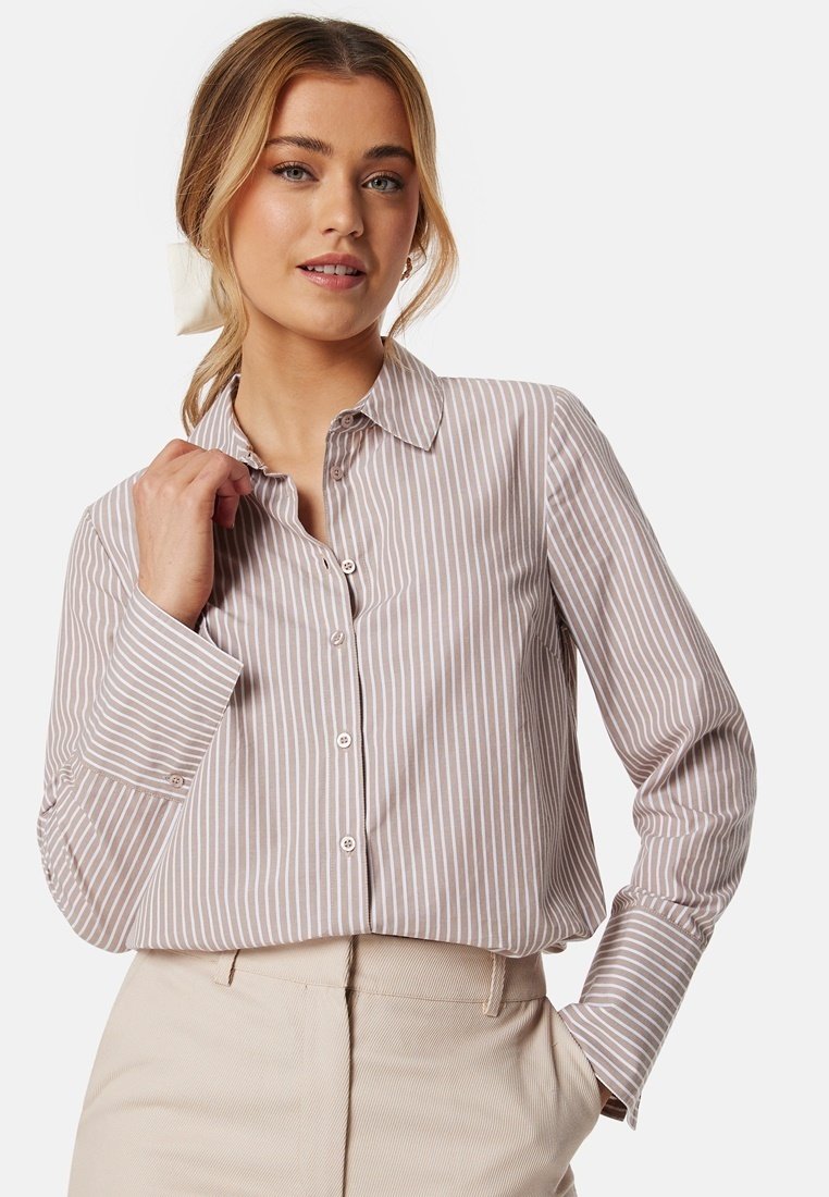 Camicia a righe color marrone chiaro e bianco con colletto, maniche lunghe e tessuto morbido, indossata con pantaloni beige a vita alta.