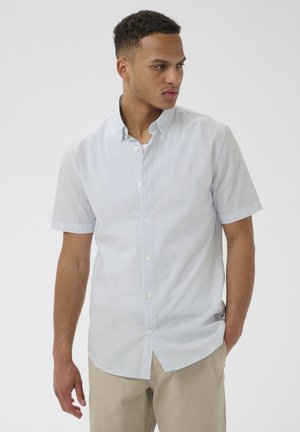 Homme regardant sur le côté, portant une chemise boutonnée à manches courtes bleu clair et un pantalon beige, sur un fond blanc uni.