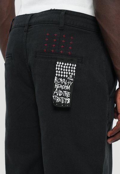Black denim pants featuring red embroidered crosses on the back and a black and white tag with text "ROYALTY HEROISM AND THE STREET."