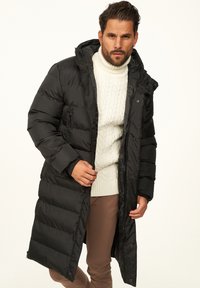 Schwarzer Steppmantel mit langem Schnitt, horizontalem Steppmuster und Kapuze. Wird über einem weißen Pullover mit Zopfstrickmuster getragen. Reißverschluss und Druckknopfverschluss.