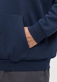 Mørkeblå hettegenser med en frontlomme og ribbestrikkede mansjetter, med teksturert stoff og avslappet passform, del av et antrekk med grå bukser.