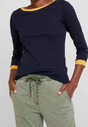 Femme portant un haut à manches longues bleu marine avec des bordures jaunes et un pantalon vert clair avec un cordon de serrage, les mains dans les poches.