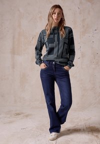 Dunkelgrüner Hoodie mit weißen geometrischen Linien, kombiniert mit dunkelblauen Jeans und weißen Sneakers. Das Modell steht mit den Händen in den Taschen.