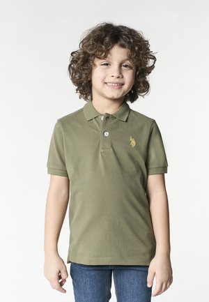 Bambino piccolo con capelli ricci che sorride, indossa una polo verde oliva e jeans blu, in piedi davanti a uno sfondo chiaro semplice.