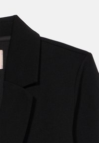 Μαύρο σακάκι με κοφτερή πέτο,  απαλό ύφασμα και δομημένους ώμους. Διακρίνεται για τη ραφή που φαίνεται και τον καθαρό, μινιμαλιστικό σχεδιασμό.