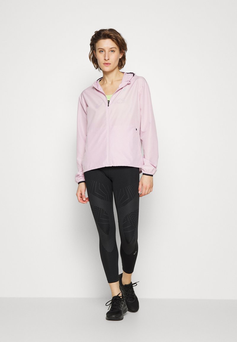 Lichtroze zip-up jack met capuchon, gecombineerd met zwarte textuurleggings met een geometrisch patroon, en zwarte sportschoenen.