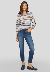 Frau in gestreiftem Langarmshirt, blauen Caprijeans und marineblauen Slippern, die mit einer Hand in der Tasche vor einem grauen Hintergrund steht.