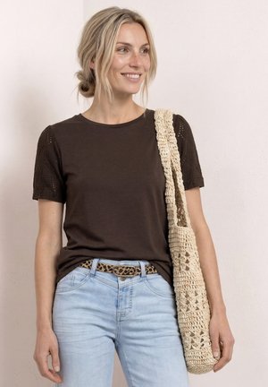Femme souriante aux cheveux blonds, portant un haut marron à manches courtes, un jean bleu clair, une ceinture à imprimé léopard et un sac à bandoulière en crochet crème.