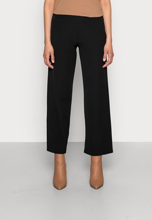 Kobieta w czarnych szerokich spodniach o długości do połowy łydki i beżowych, spiczastych botkach za kostkę, stojąca na tle jasnej, gładkiej ściany.